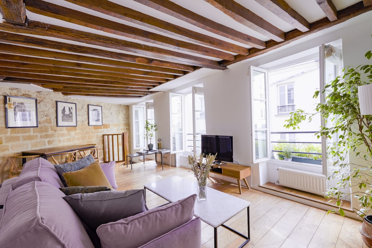 A spacious living area features exposed wooden beams and light-colored walls. Large windows open to a balcony, allowing natural light to fill the room. A soft purple sofa, minimalist coffee table, and decorative plants create a relaxed environment, while a television stands nearby.
