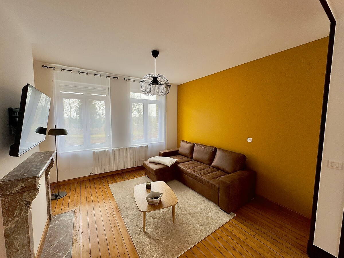 A bright living area features two large windows allowing natural light to fill the space. A brown sectional sofa sits adjacent to a modern coffee table, with a flat-screen TV mounted on the wall opposite. Light-colored wood flooring complements the warm accent wall.