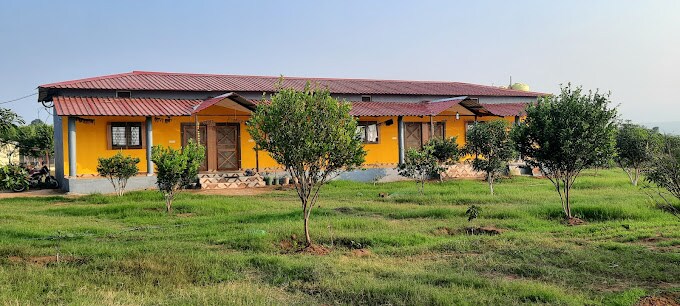 A single-story building with a vibrant yellow facade is complemented by a red metal roof. The structure is flanked by small trees and greenery, set against an expansive lawn that invites outdoor activities. Window shutters are closed, providing a serene atmosphere.
