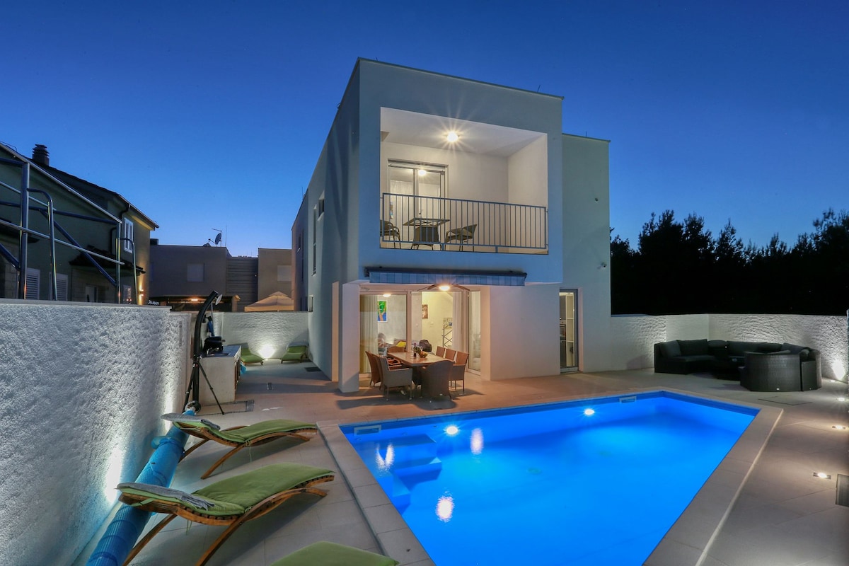 A modern villa is shown at dusk, featuring a bright blue swimming pool surrounded by lounge chairs. The spacious outdoor area includes a patio with seating, and large glass doors lead into the interior, highlighting a blend of indoor and outdoor living.