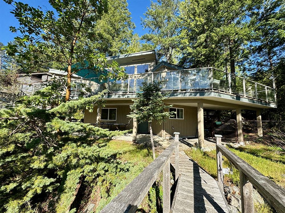 The two-story house is surrounded by trees, featuring a spacious wraparound balcony. A wooden pathway leads to the entrance, inviting guests to explore the outdoor space. The balcony is enclosed by glass railings, providing unobstructed views of the natural surroundings.