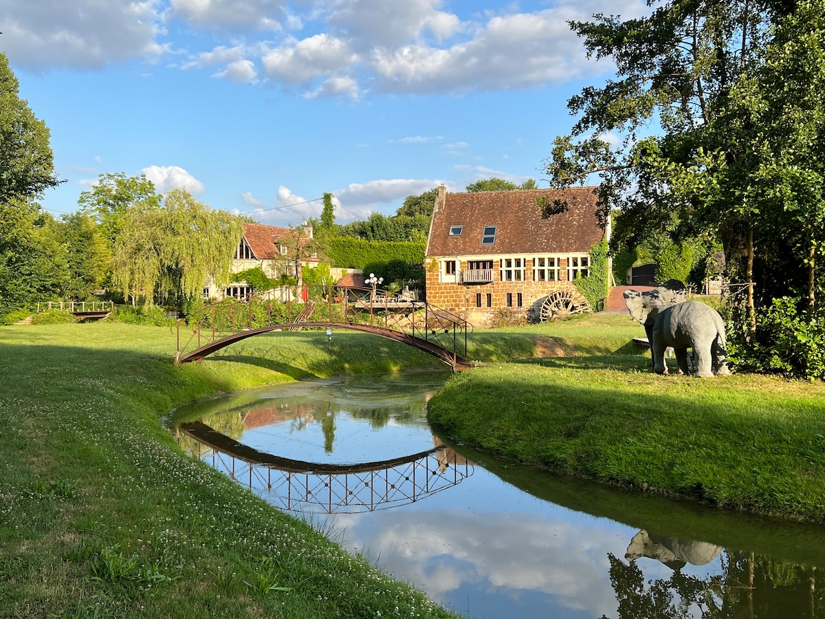 A serene landscape is captured, featuring a charming stone building beside a tranquil pond. A small bridge arches over the water, reflecting greenery and clouds above. Sculptures, including a bear, can be seen amidst the lush grass, enhancing the natural setting.