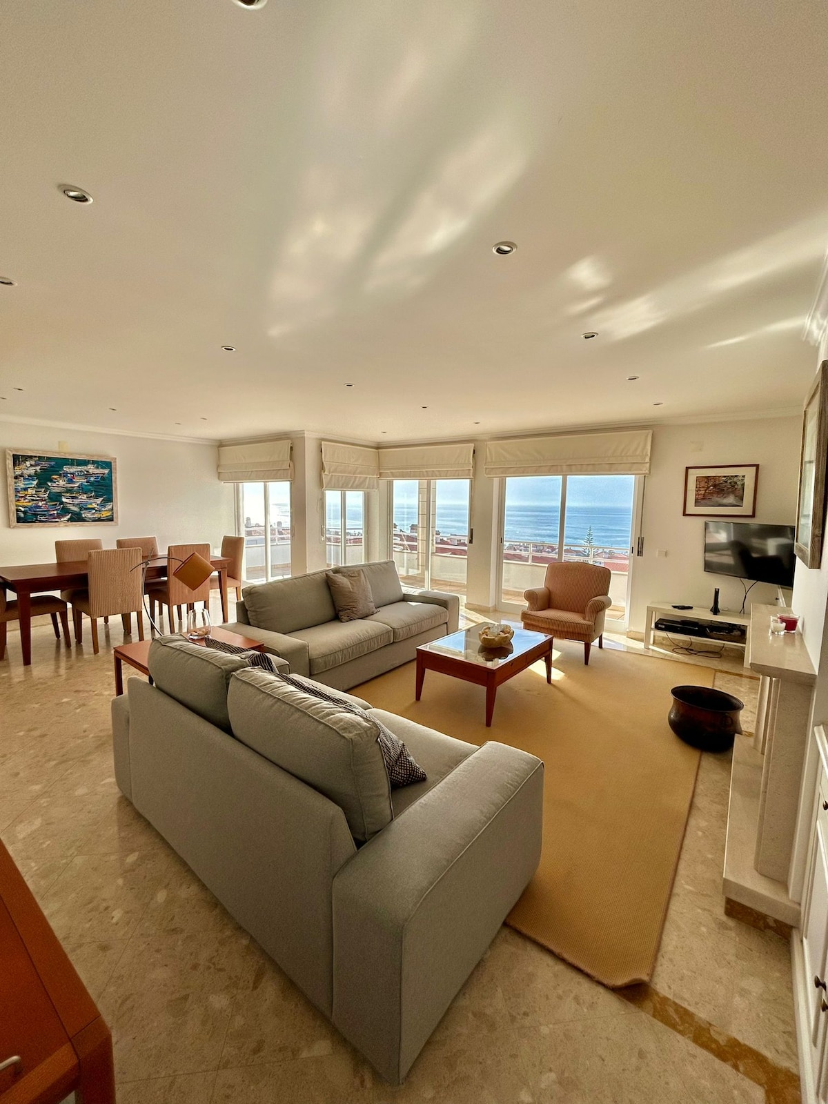 A well-lit living area features comfortable seating arrangements, including two gray sofas and a wooden coffee table. Large windows provide views of the sea and allow natural light to fill the space. A dining area is visible in the background, enhancing the open layout.