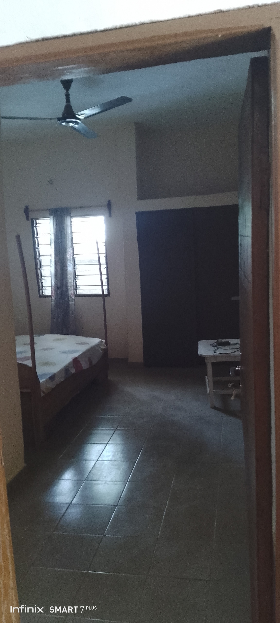 A spacious room is visible, featuring a large bed with a light-colored bedspread. A ceiling fan can be seen overhead. Windows with partially open shutters allow natural light to enter, illuminating the tiled flooring. A wardrobe and a small table are also present.