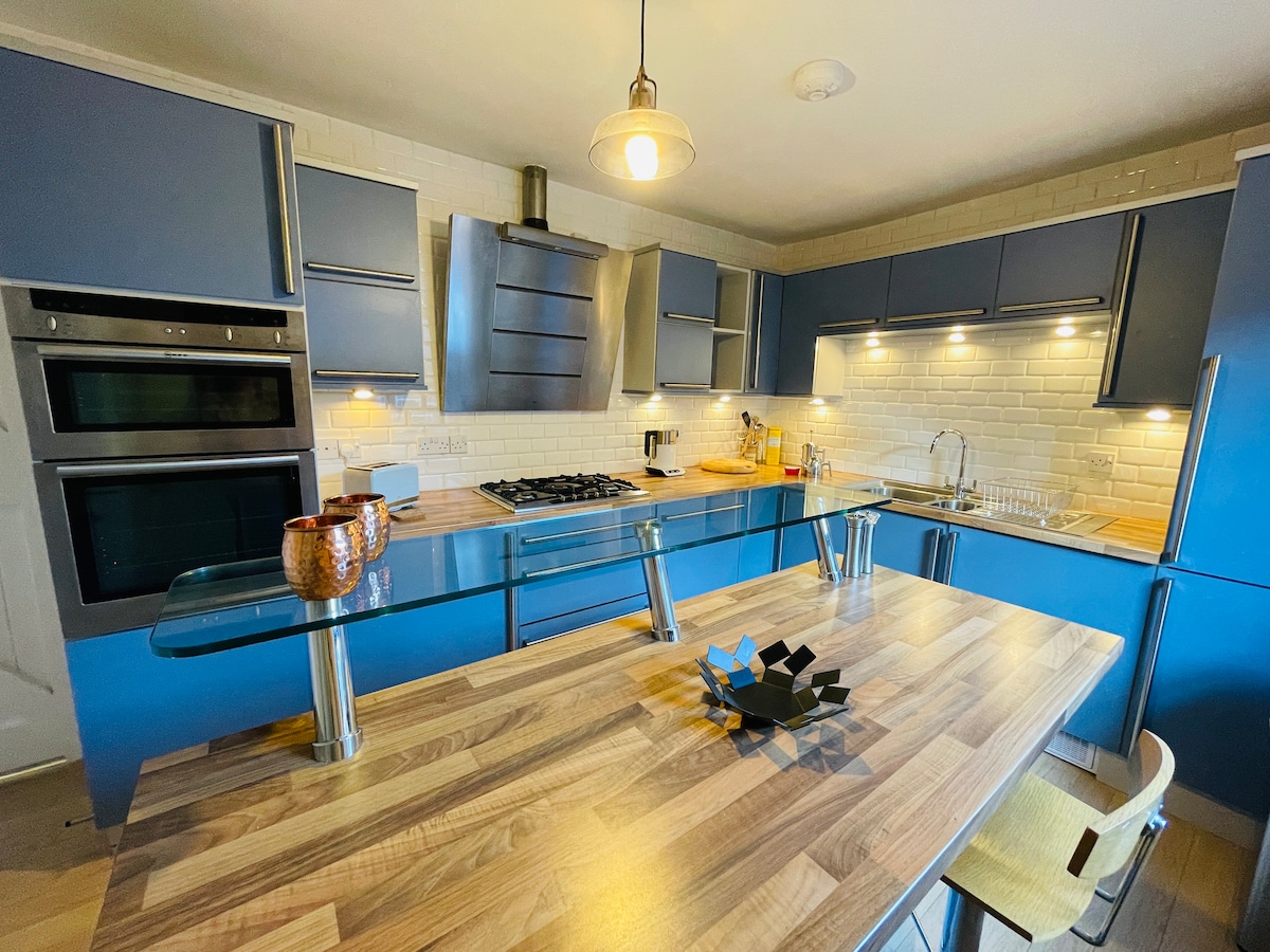 A modern kitchen features sleek blue cabinetry paired with wood accents. Stainless steel appliances, including a gas stove, are mounted against a textured backsplash. A glass-topped island is accompanied by contemporary seating and a decorative centerpiece, enhancing the functional layout of the space.