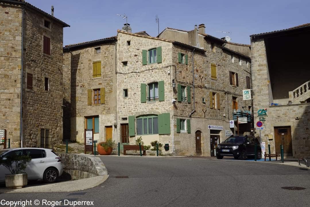 The image depicts a charming stone village building at a street corner, featuring varied window shutters in green and yellow. A parked car is visible nearby, along with potted plants at the base of the building. The scene reflects the rustic architecture of the medieval town.