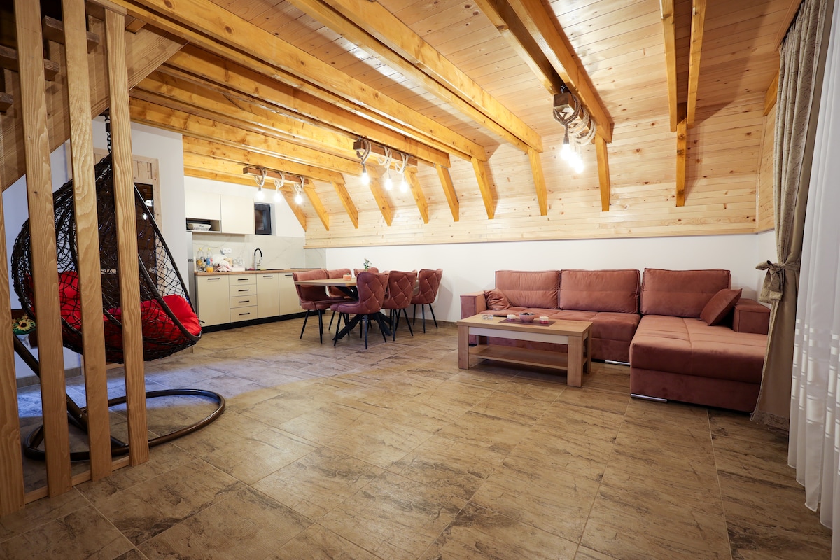 The interior features a spacious living area with wooden beams overhead. A sectional sofa is positioned alongside a coffee table, while a dining area with elegant chairs is visible. A woven hanging chair adds a unique touch to the room's design.