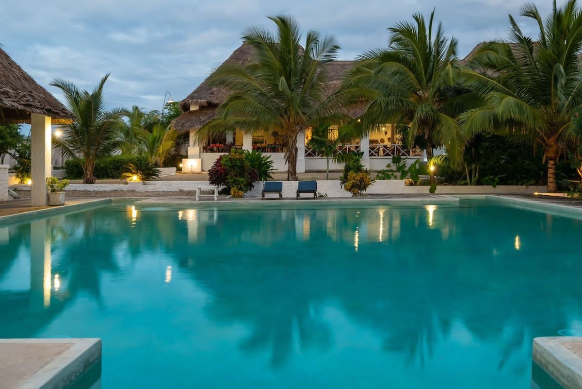 A large swimming pool reflects the surrounding palm trees and the serene sky, creating a calm atmosphere. Comfortable seating areas are positioned nearby, and the main building features thatched roofing with outdoor dining options visible.