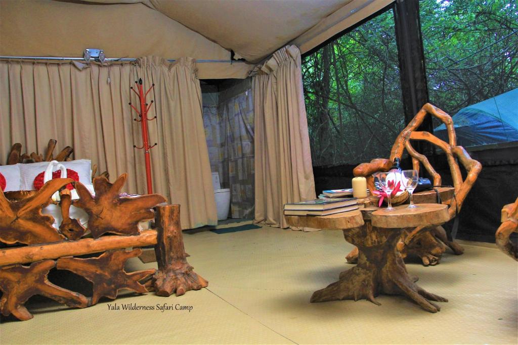 The interior space features natural wooden furniture, including a unique coffee table and a comfortable seating area. Beige curtains frame the entrance, and the flooring is light-colored. A small bookshelf showcases items, while a nearby area offers visibility of camping essentials.