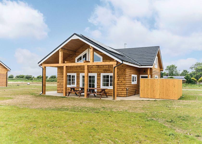 A spacious wooden cabin is depicted, featuring a prominent covered porch with picnic tables. Large windows allow natural light to fill the interior, while the surrounding grassy area enhances the serene countryside setting.