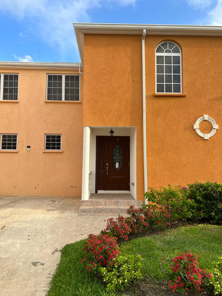 The entrance of the property features a warm orange exterior with large windows. A framed door, adorned with glass accents, is positioned centrally, while vibrant flowers line the walkway, adding touches of color. A well-manicured lawn enhances the welcoming appearance.