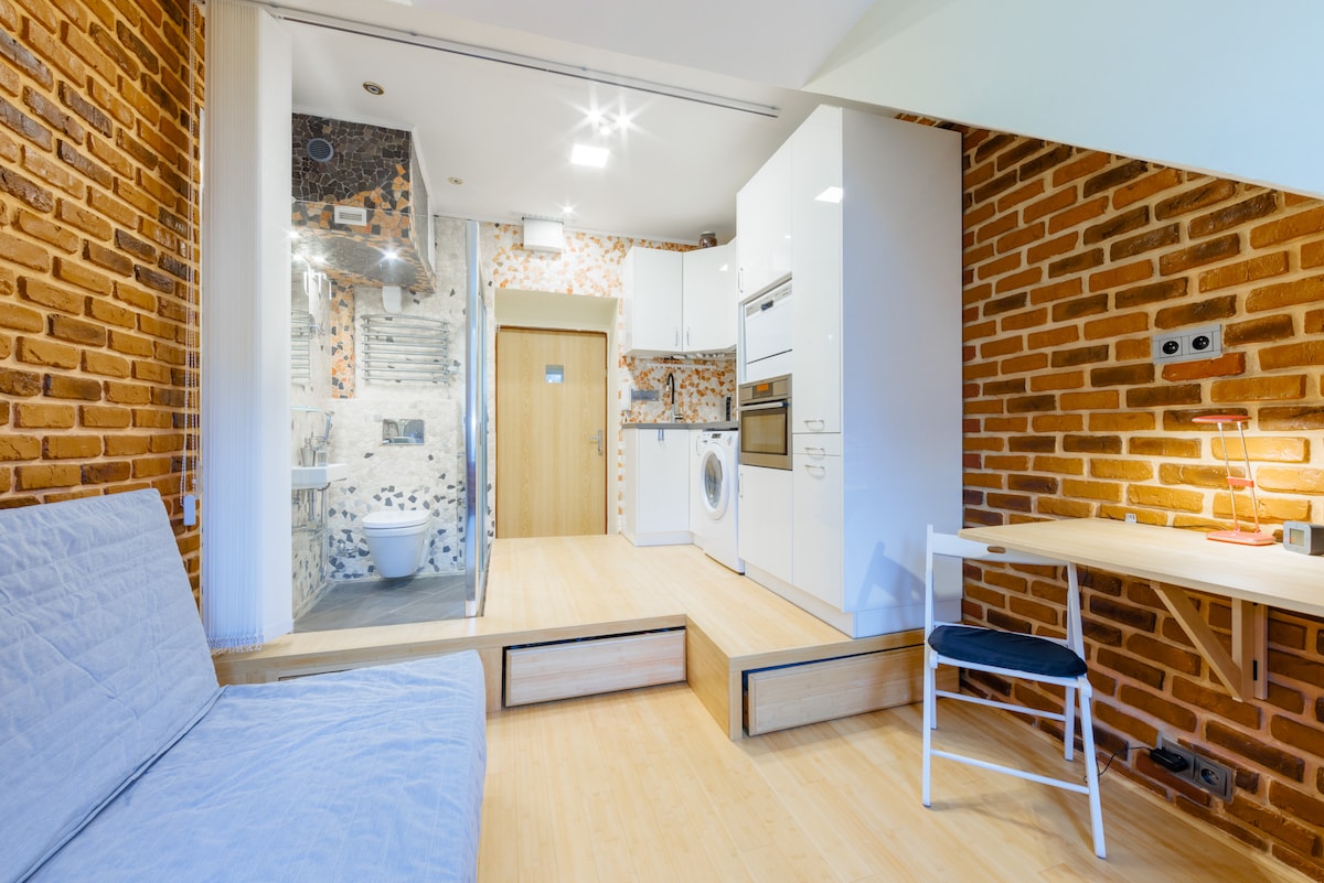 The studio apartment features a modern kitchen with white cabinetry and a washing machine. A cozy seating area includes a sofa bed, while light wooden flooring enhances the space. Exposed brick walls add texture, and a small desk accompanies a chair, creating an inviting workspace.