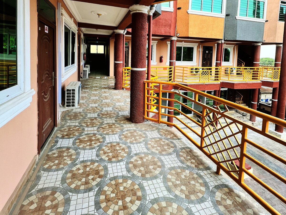 A tiled walkway presents a colorful geometric pattern, bordered by railings and supported by sturdy pillars. Air conditioning units are visible along the walls, contributing to a comfortable environment. The corridor leads to elaborate spaces designed for guest access, with natural light enhancing the overall ambiance.