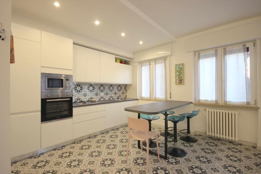 A modern kitchen features white cabinetry, a patterned tile floor, and a central table surrounded by four sleek chairs. Natural light filters through multiple windows, illuminating the space. Appliances include a built-in oven and a microwave, enhancing functionality.