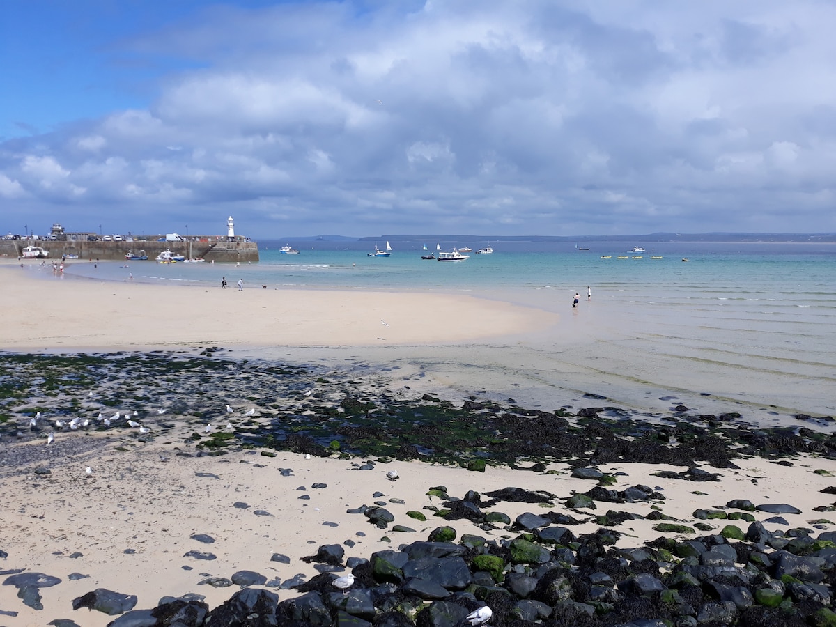 A sandy beach stretches into the distance, bordered by calm turquoise waters. In the foreground, dark stones and patches of green seaweed are visible along the shoreline. Small boats can be seen in the background, while a lighthouse stands at the edge of the harbor under a partly cloudy sky.