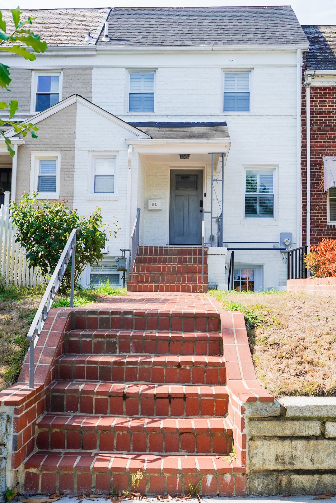 The entrance to the fully renovated apartment is marked by a set of red brick stairs leading to a gray door. The white exterior of the house contrasts with the surrounding greenery, while a grassy area is visible beside the stairs.