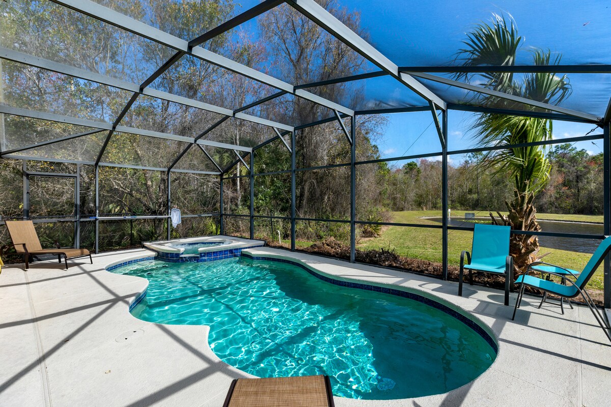 A private screened swimming pool and spa are showcased, surrounded by lush greenery. Comfortable loungers are positioned for sunbathing, while a vibrant blue chair offers an additional space for relaxation. The pool features a unique shape, providing an inviting atmosphere for leisure activities.
