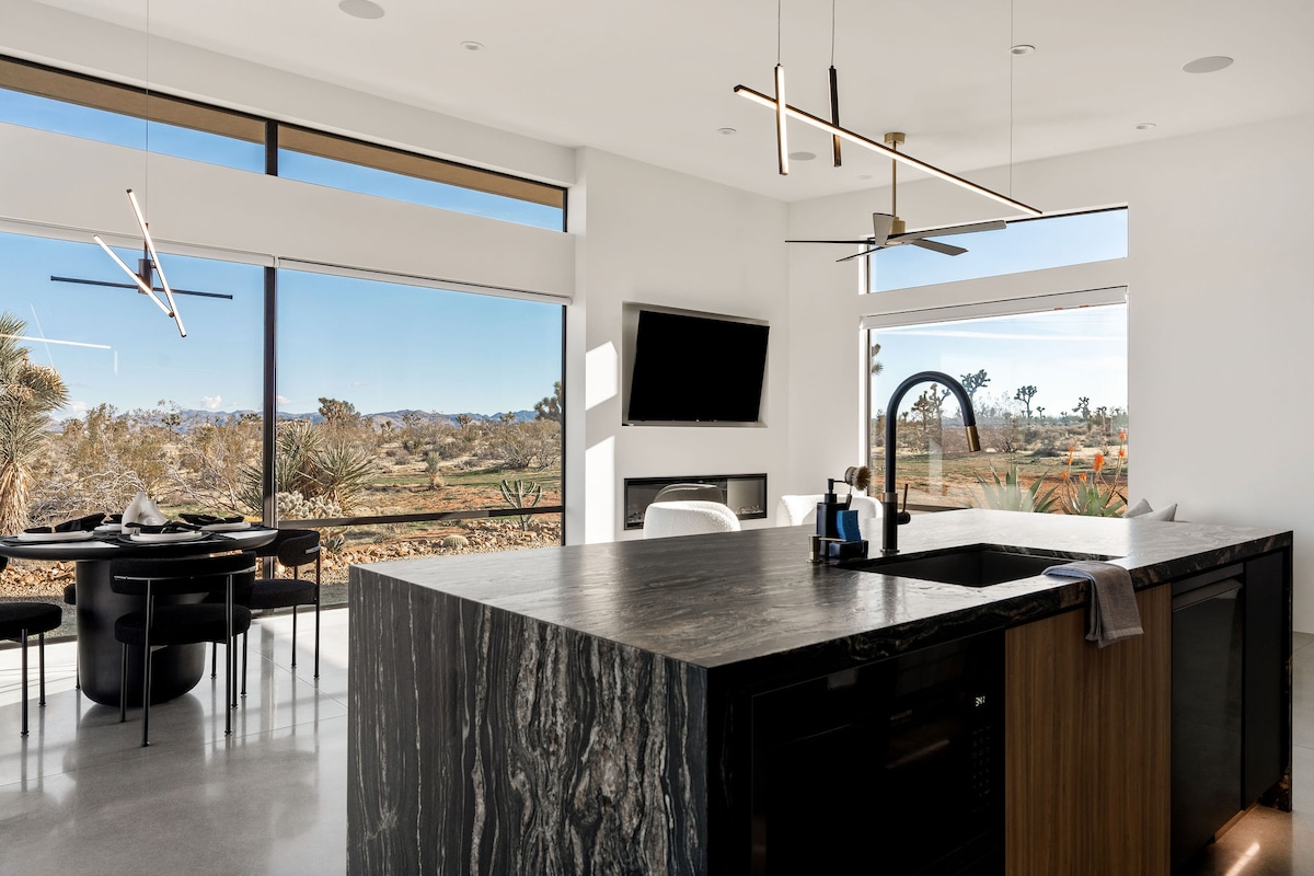 A modern kitchen and dining area features a large island with dark, textured countertops and ample seating. Expansive windows provide scenic desert views, while sleek lighting fixtures and a television enhance the contemporary design. A cozy dining table is positioned nearby for enjoyable meals.