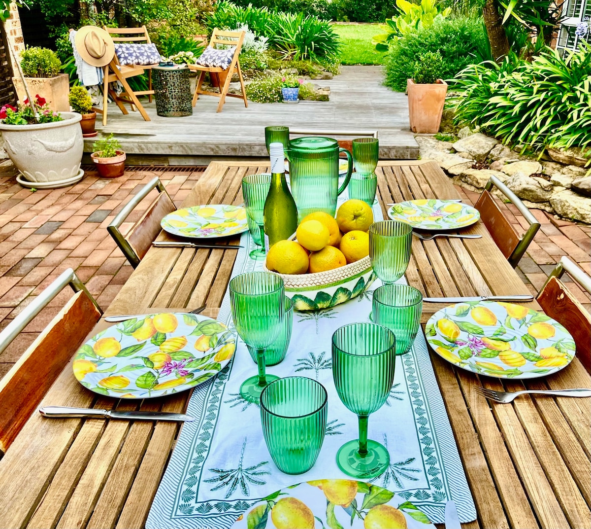 A wooden dining table is set with green glassware and plates featuring a lemon design. In the center, a pitcher and bowl of lemons are displayed. The background includes a patio area with green foliage, wooden chairs, and decorative pots, creating a lively outdoor dining space.