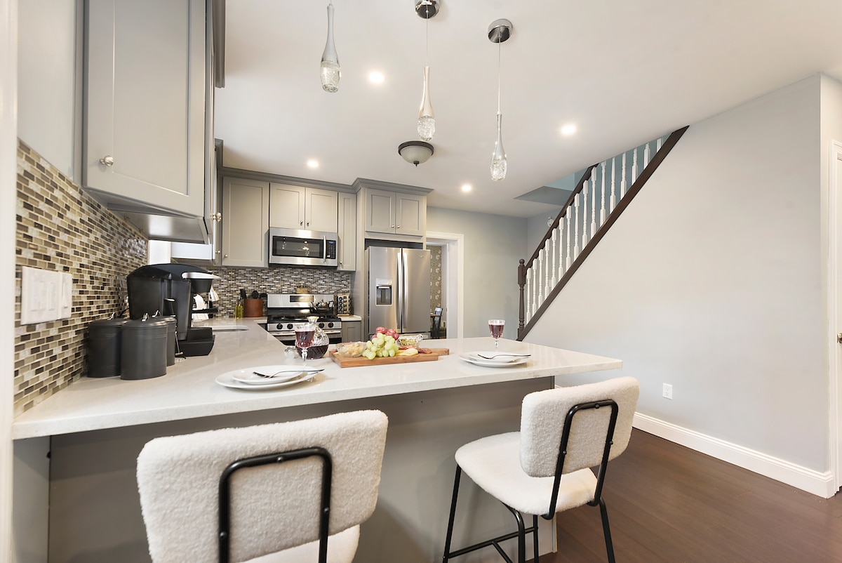 A modern kitchen features a breakfast bar with two upholstered stools. Light fixtures hang above the counter, which is adorned with a fruit platter. Stainless steel appliances, a dark backsplash, and light cabinetry create a sleek, inviting cooking space.