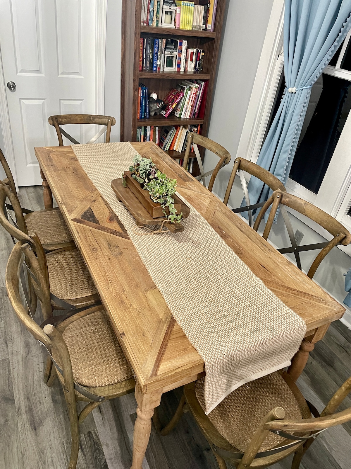 A wooden dining table is surrounded by six chairs with woven seats. A neutral-toned table runner is placed at the center, adorned with a decorative planter. A bookshelf featuring various books is partially visible in the background, alongside soft curtains on the window.