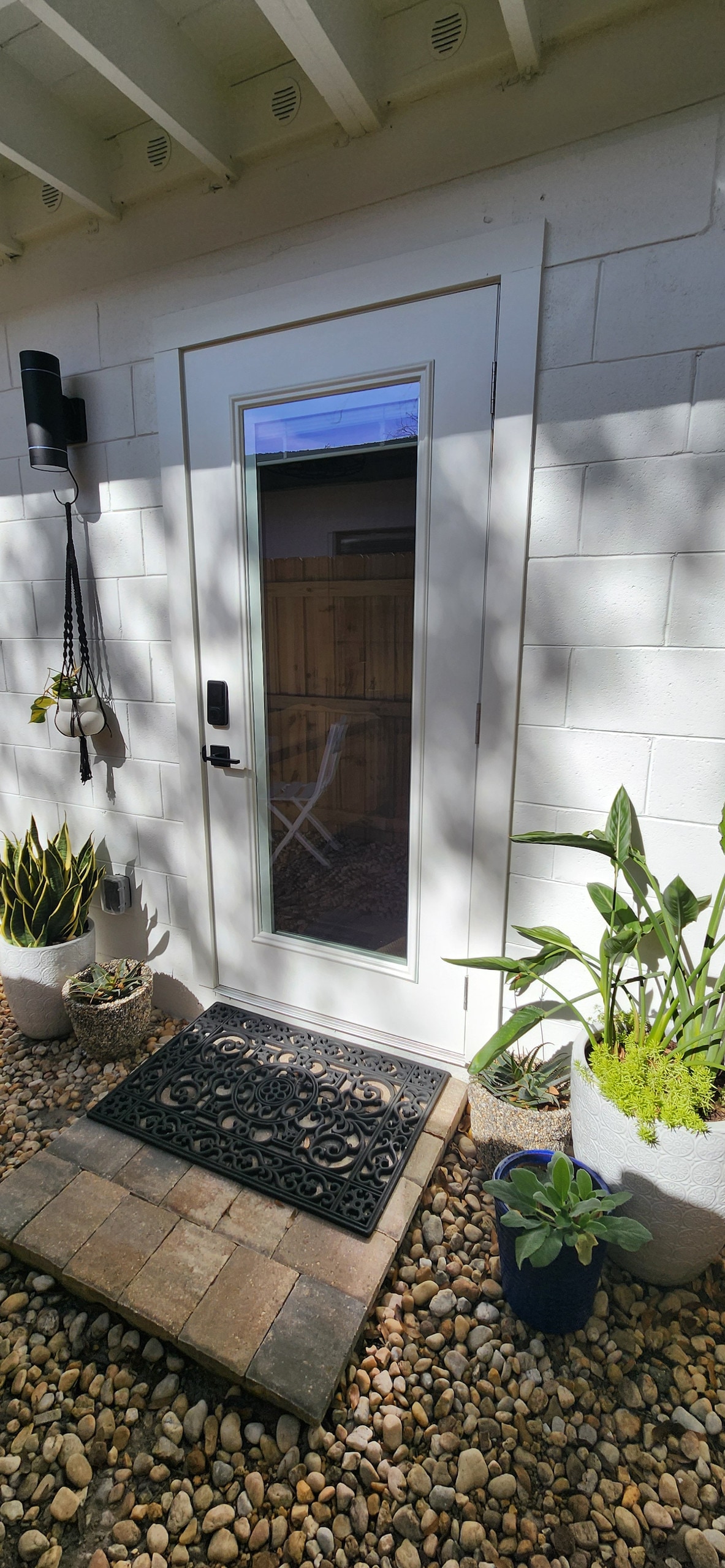 A white door with a glass panel leads into the studio, framed by a decorative welcome mat on stone pavers. Lush green plants are positioned beside the door, adding a natural touch to the entrance. Soft shadows from nearby foliage create a calming atmosphere.