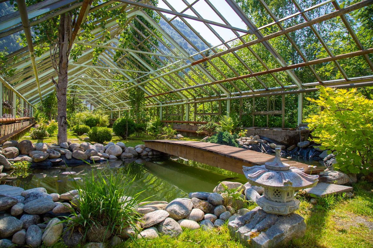 An indoor garden area features a pond surrounded by smooth stones, with greenery visible throughout the structure. A wooden bridge crosses over the pond, and a decorative stone lantern is positioned nearby, adding a calming ambiance to the space.