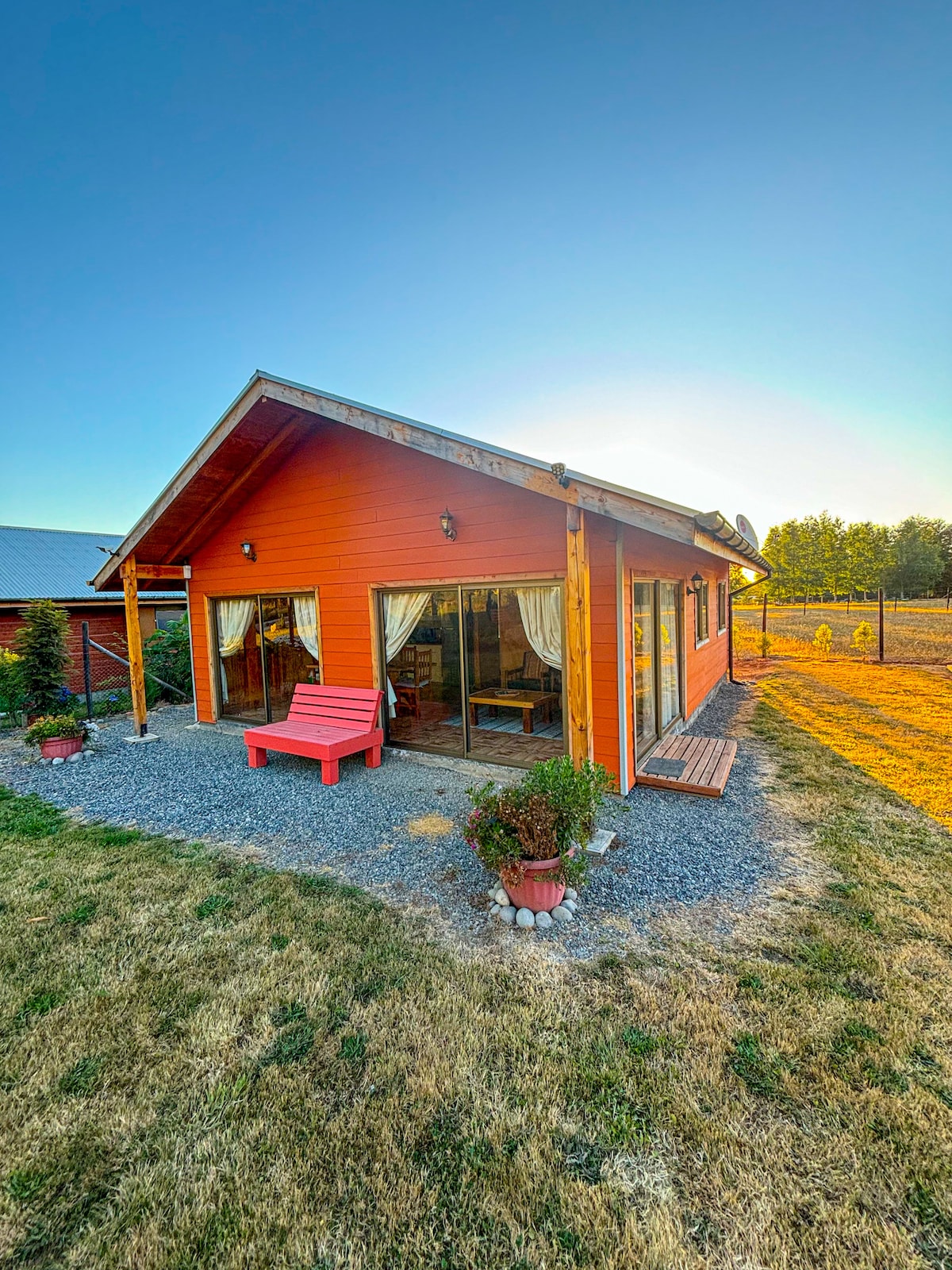 An orange wooden house is presented, featuring large glass doors and a welcoming porch area with seating. The surrounding landscape is open, including a grassy area and flower pots. Sunlight highlights the exterior, creating a warm atmosphere in the late afternoon.