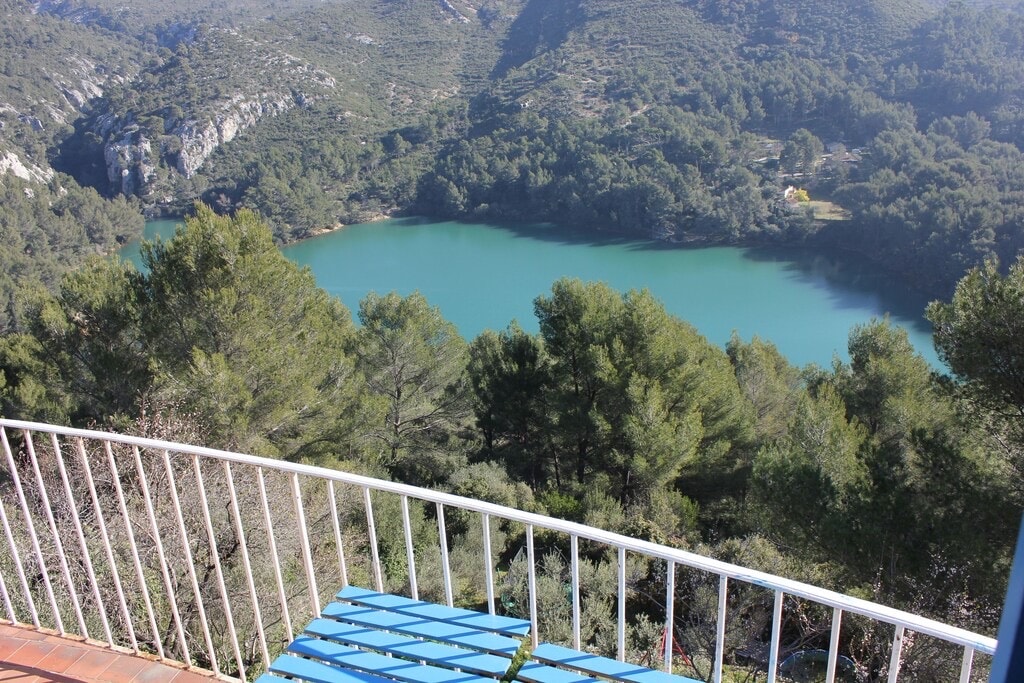 A tranquil view showcases a serene turquoise lake surrounded by lush greenery and hills. A balcony railing frames the landscape, with a blue patio chair inviting relaxation while enjoying the natural scenery in the distance.