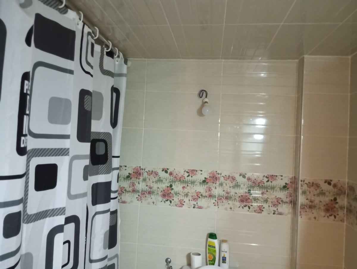A bathroom setting is depicted with tiled walls featuring a floral accent strip. A patterned shower curtain is visible, and personal care items are neatly arranged on the ledge. Soft lighting highlights the clean and functional design of the space.