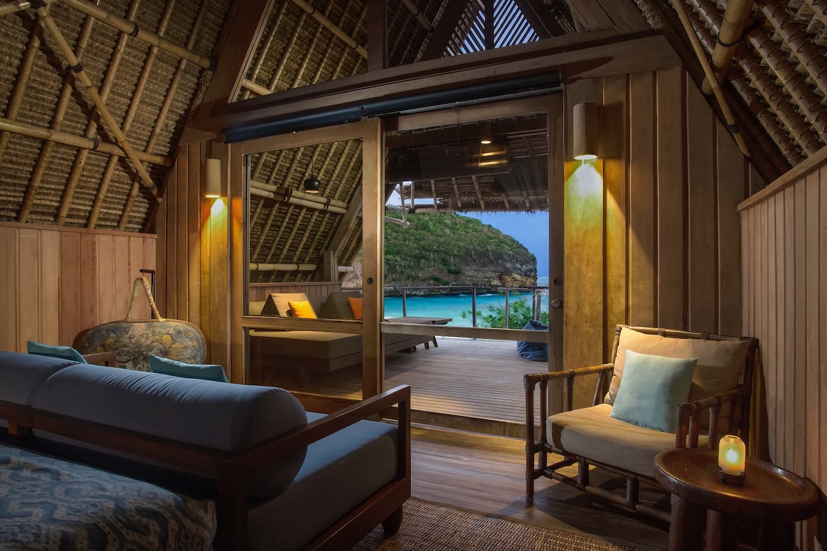 An inviting interior features wooden walls and a high ceiling with bamboo accents. A comfortable seating area, complete with a sofa and armchairs, overlooks a private balcony. Soft lighting enhances the cozy atmosphere, while ocean views are visible through glass doors.