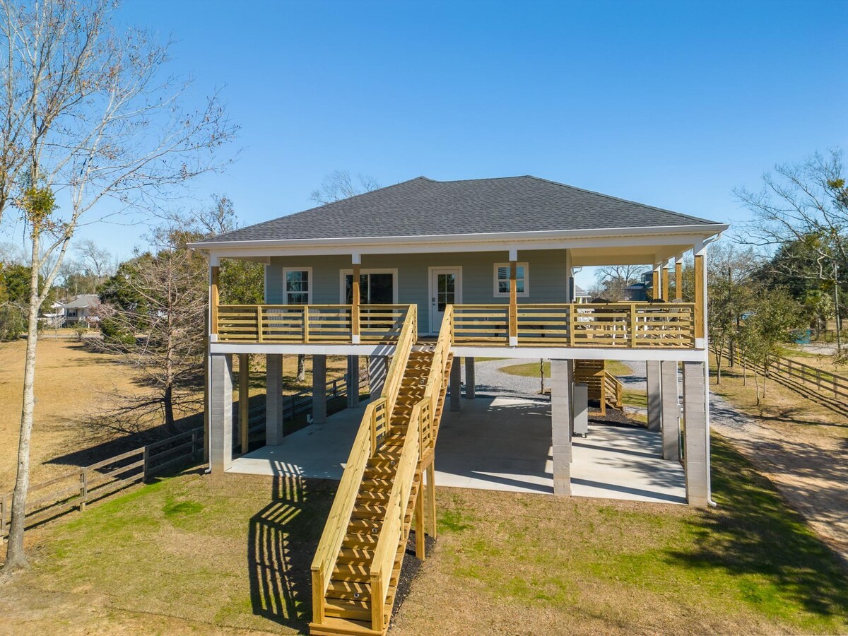 A newly built two-story property is shown with a spacious deck featuring wooden railings and stairs leading to the ground level. The exterior is neutral-toned, set against a clear blue sky. The surrounding landscape includes open grassy areas and scattered trees.