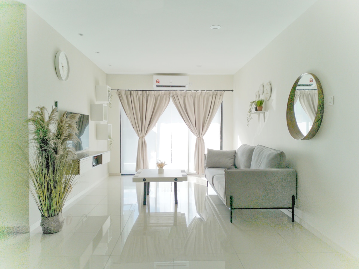 A bright living area features a gray sofa positioned beside a minimalist coffee table. Natural light floods the space through large glass doors, with sheer curtains gently framing the view. A round mirror and decorative wall accents add subtle details, complemented by a potted plant.