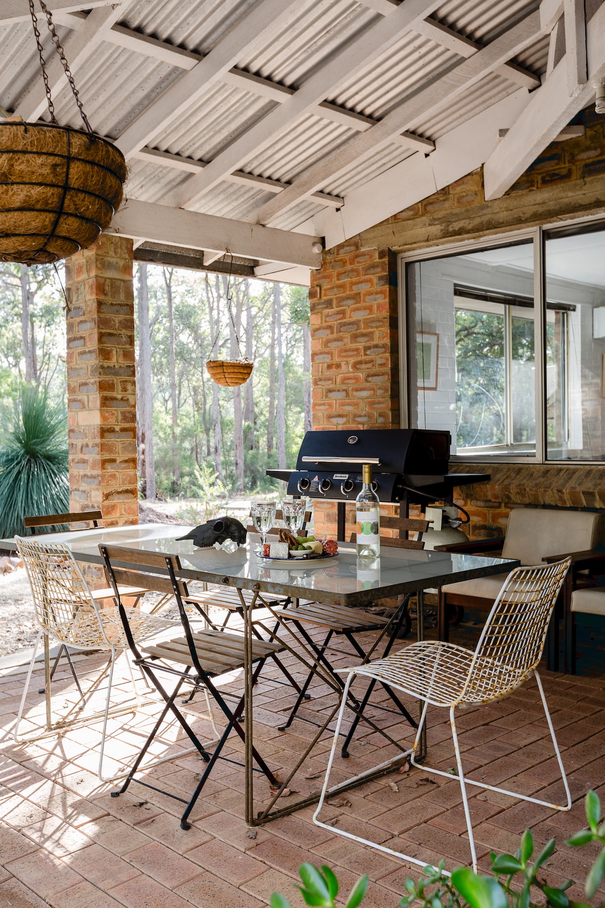 A covered outdoor patio area is presented, featuring a metal dining table surrounded by four wire chairs. A barbecue grill is positioned nearby, with natural surroundings visible through large windows. Decorative hanging baskets add a touch of charm to the rustic setting.