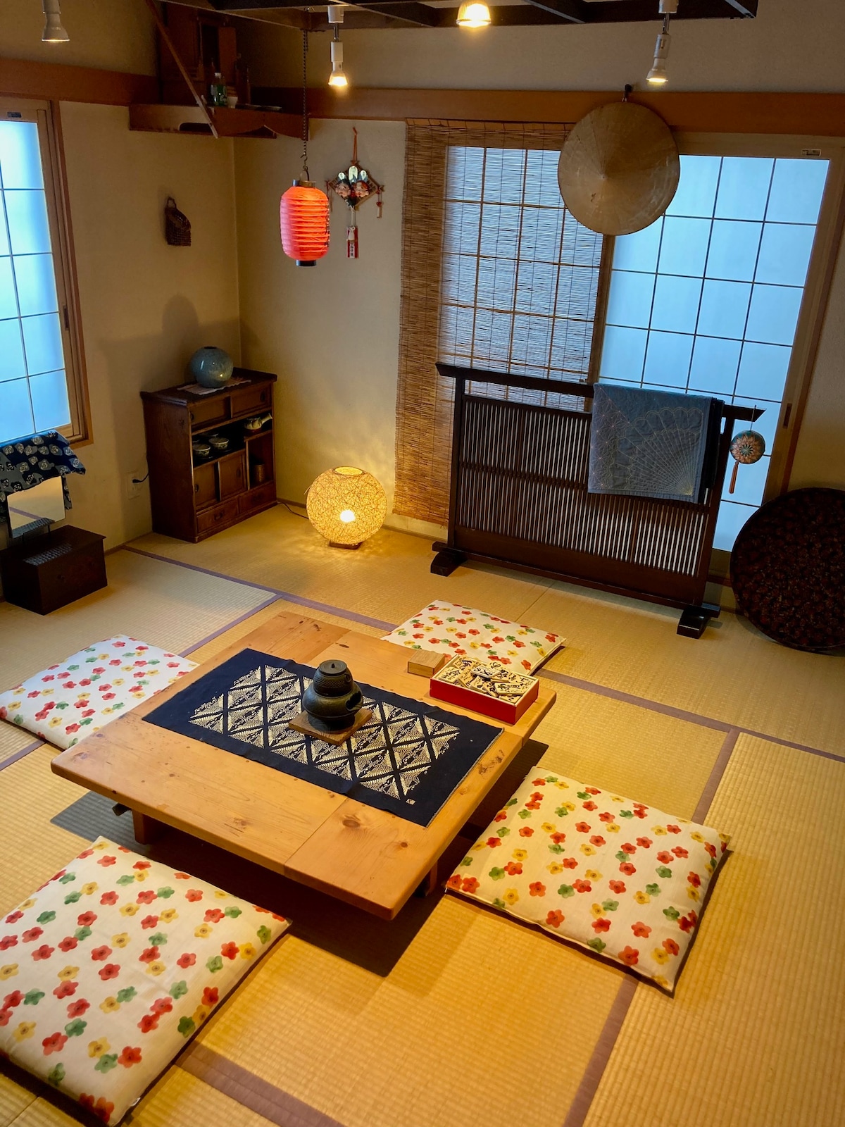 A traditional Japanese tatami room is showcased with a low wooden table at the center, surrounded by floor cushions featuring floral patterns. Soft lighting illuminates the space, while shoji screens and decorative elements lend a serene ambiance.