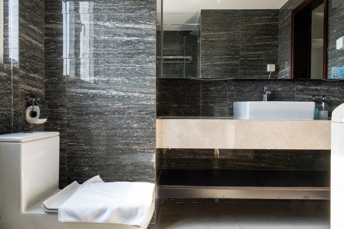 A modern bathroom features sleek dark stone walls and a spacious vanity with a large mirror. A white sink is positioned above a light-colored countertop. A white towel is neatly placed on the toilet seat, while a glass shower is visible in the background.