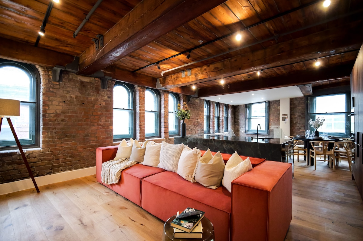 A spacious living area features a contemporary orange sectional sofa, complemented by decorative pillows. Exposed brick walls and wooden beams add character, while large windows allow natural light to fill the space. A dining area and modern kitchen can be seen in the background.