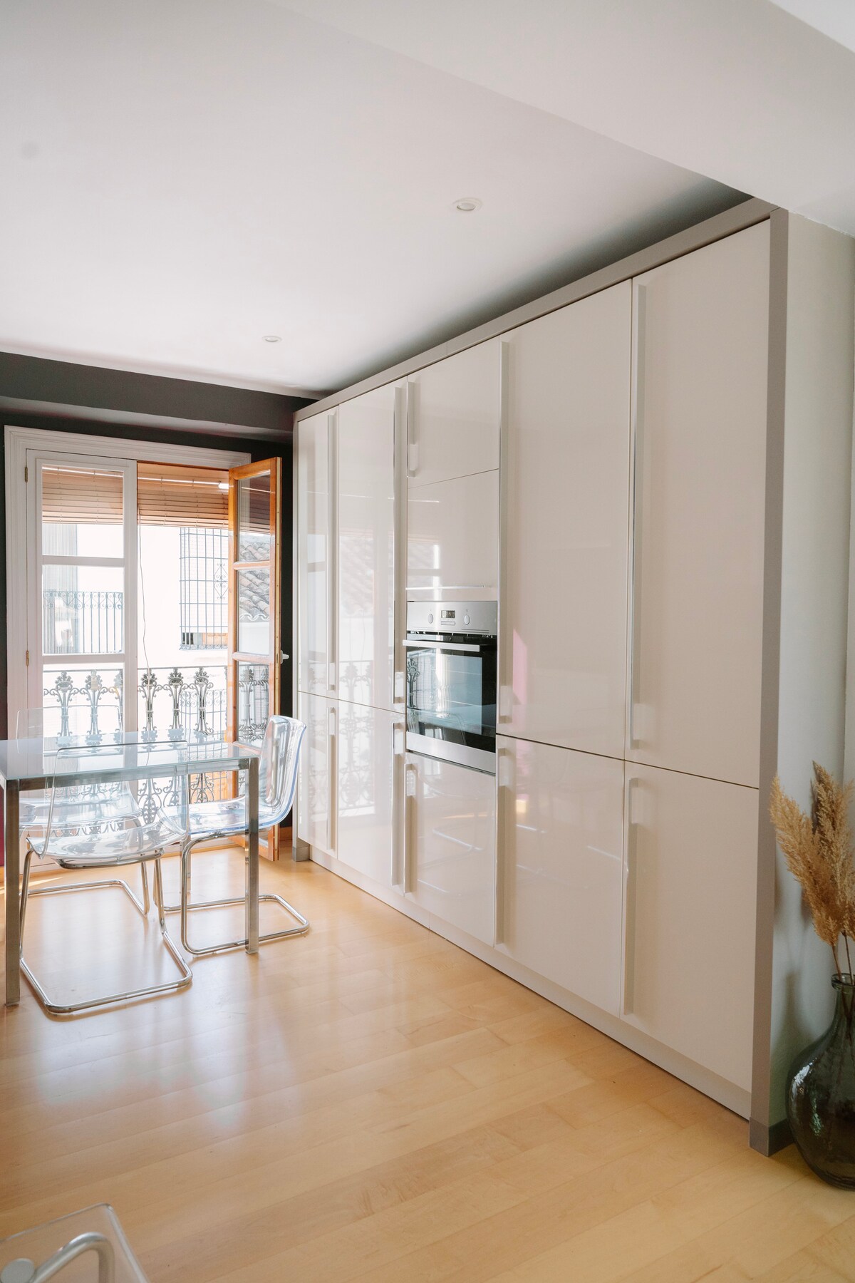 A modern kitchen features sleek, high-gloss cabinetry paired with a minimalist dining table and transparent chairs. Large windows allow natural light to fill the space, with a view of the balcony railing and cityscape beyond.