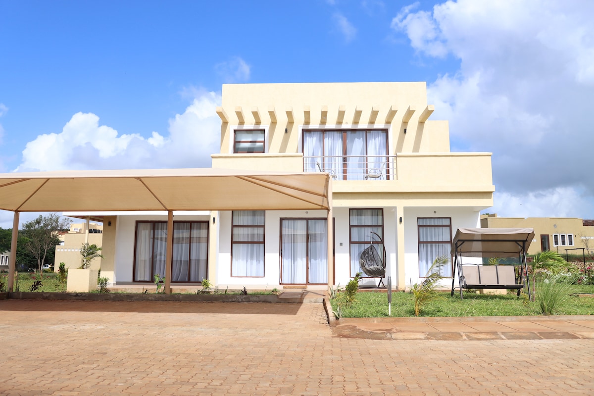 A modern villa is showcased with a spacious front porch. The structure features large windows that flood the interior with natural light, and a stylish overhang provides shade. The surrounding landscape includes green plants and a neatly maintained pathway.