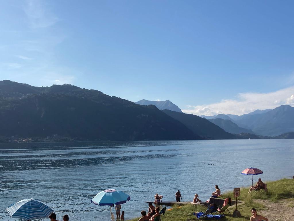 A serene lakeside scene displays calm water reflecting the clear sky, with mountains rising in the background. Colorful umbrellas provide shade as visitors relax on the grassy shore, enjoying the tranquil environment and pleasant weather.