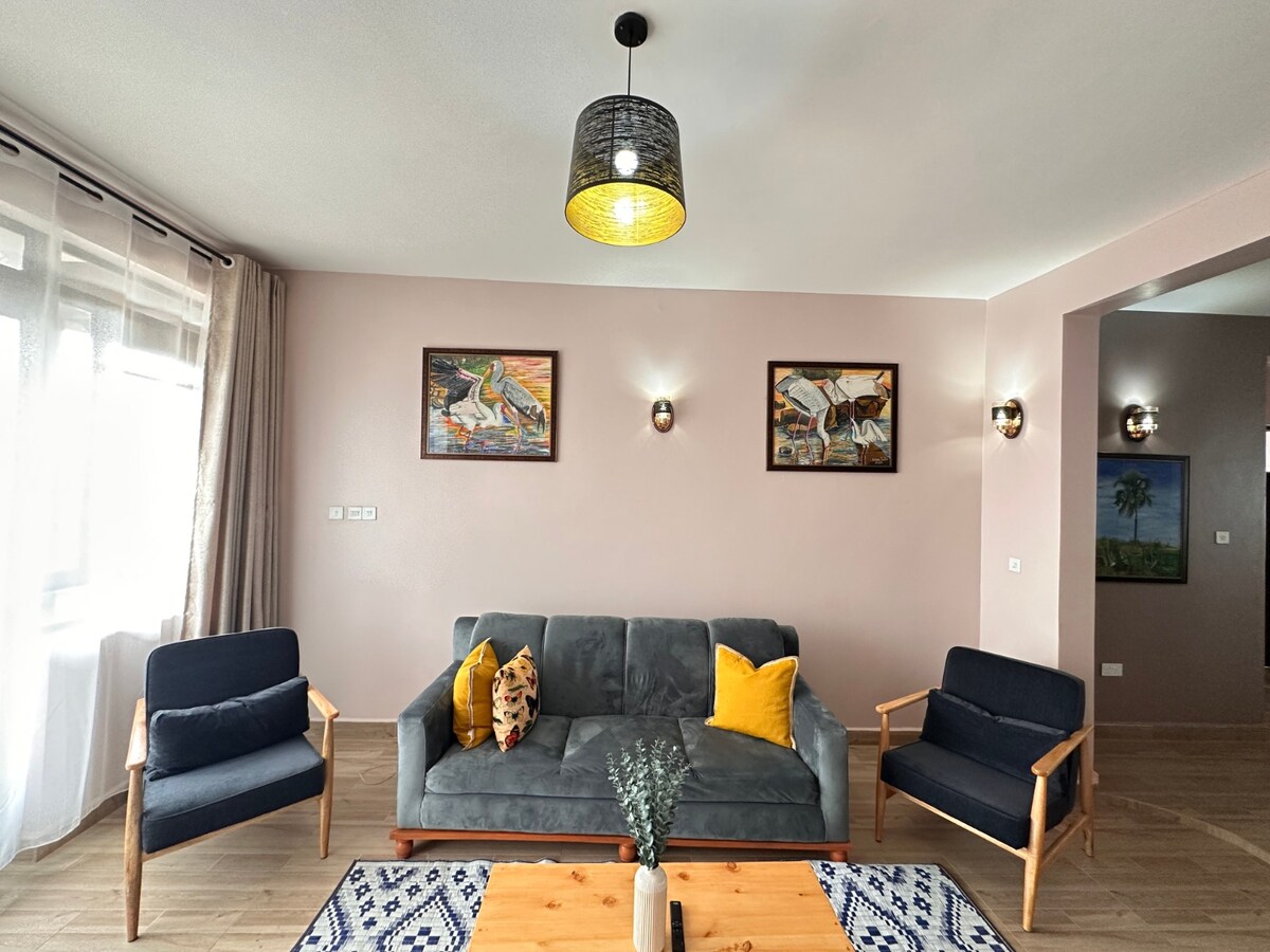 A cozy living area is adorned with two framed artworks mounted on light-colored walls. A spacious gray sofa with yellow and patterned cushions is complemented by two wooden armchairs. Natural light floods the space through large windows, enhancing its inviting atmosphere.