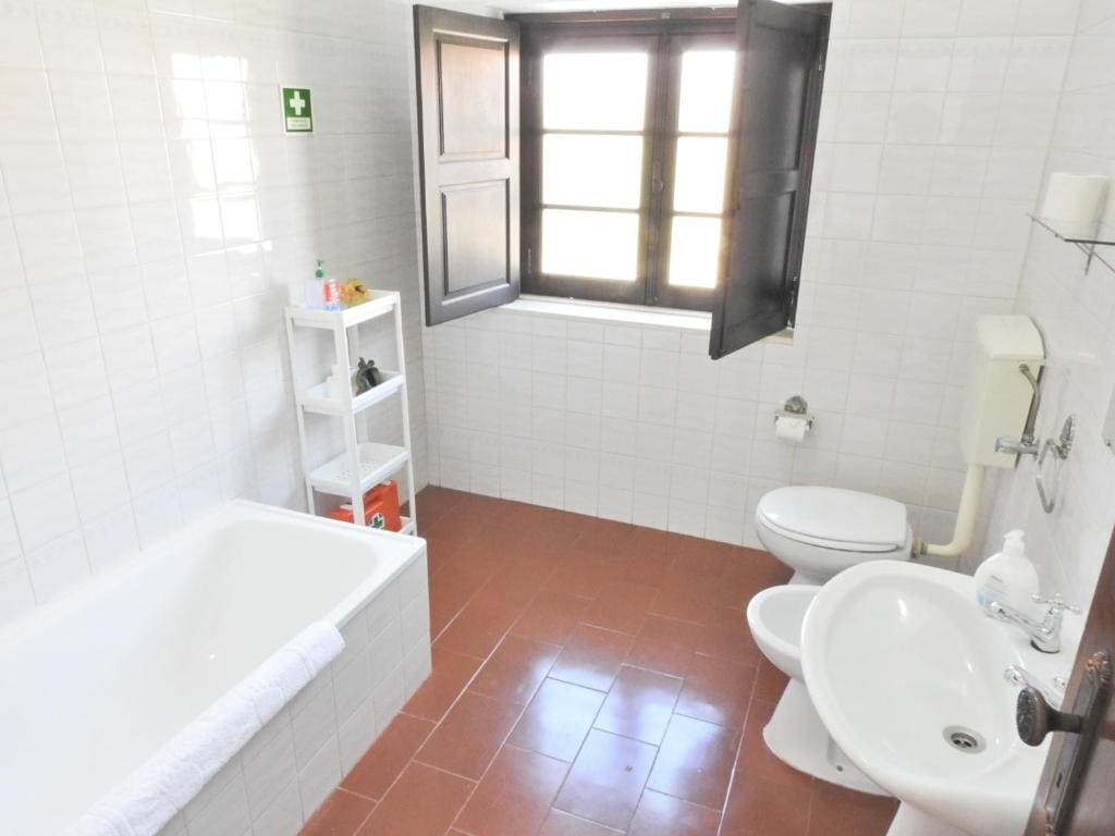 A private bathroom features a bathtub beside a window, allowing natural light to enter. A white shelving unit displays toiletries, while a toilet and sink complete the space. The light-colored tiles and warm terracotta floor create a clean and welcoming ambiance.