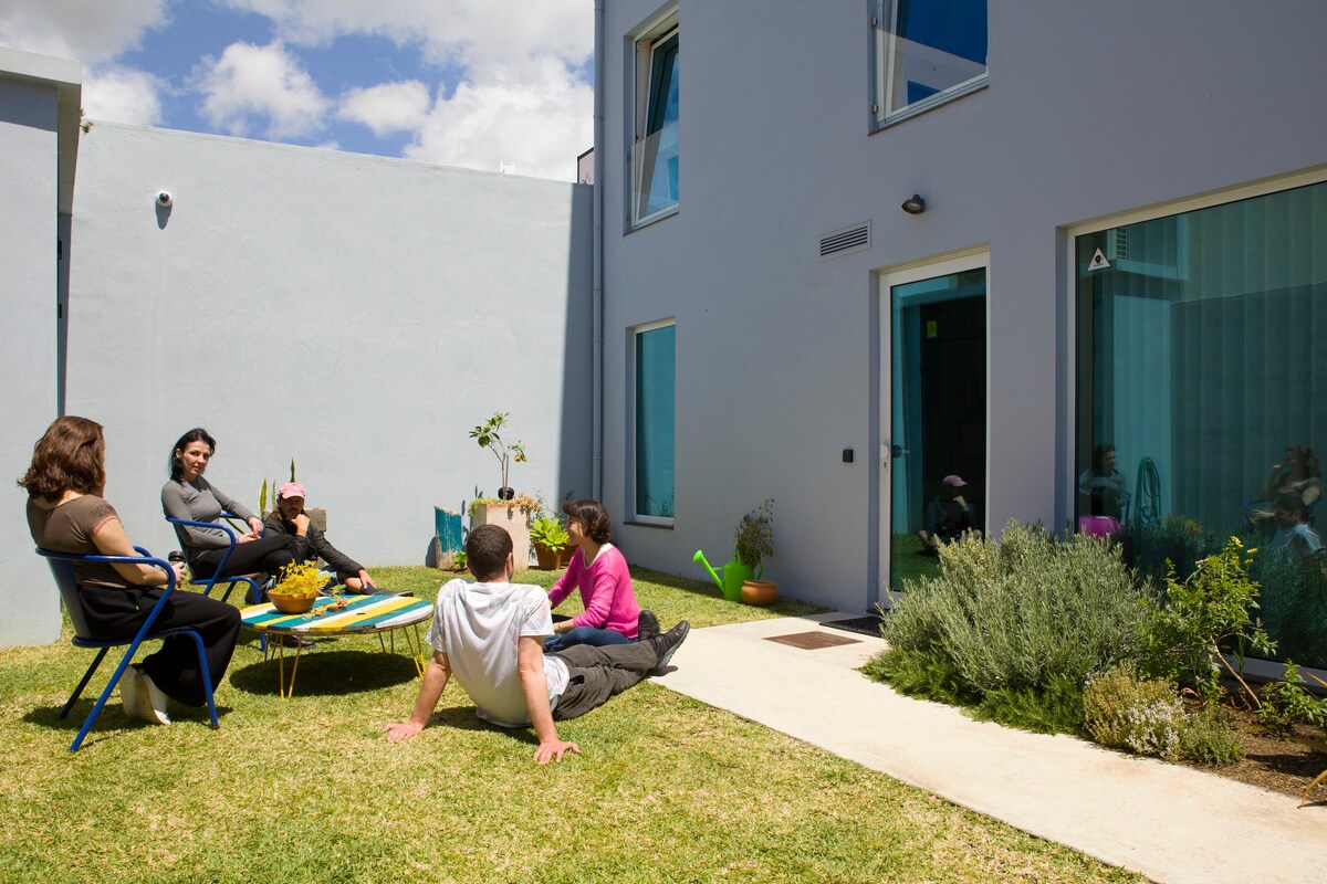 A designed outdoor space is visible, featuring a grassy area with a few chairs and a low table. The light-colored building has large windows, emphasizing natural light. Potted plants and a relaxing atmosphere invite gatherings in this shared area.