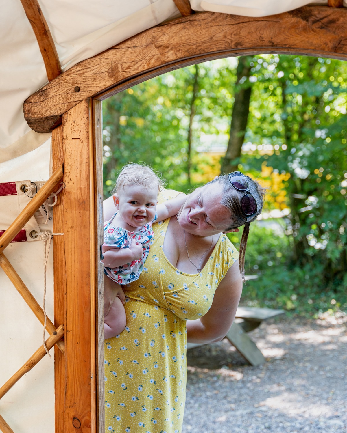A doorway of the yurt opens into a sunlit outdoor area, framed by wooden accents. A parent with a child on their back is seen against a backdrop of lush green foliage. Outdoor seating is indicated in the background, enhancing the inviting atmosphere.