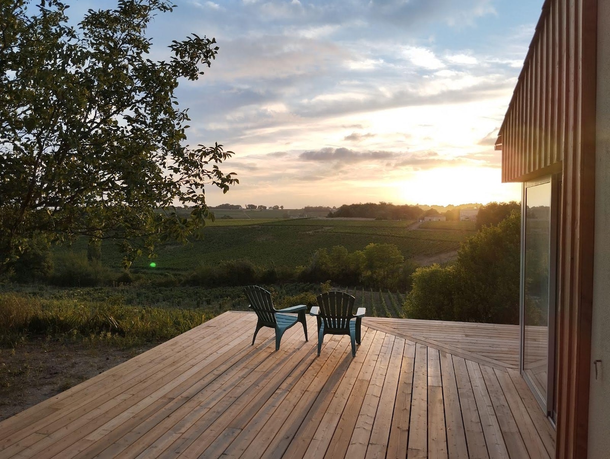 A spacious wooden terrace is shown, featuring two green Adirondack chairs facing a scenic view of vineyards and rolling hills. The sky is illuminated by a warm sunset, creating a serene atmosphere, with gentle greenery surrounding the area.