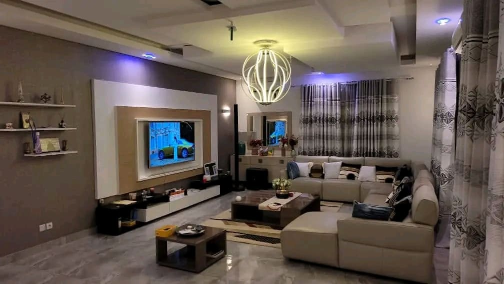 A spacious living area is equipped with a large sectional sofa, facing a wall-mounted television. Soft lighting is provided by a modern pendant fixture. Decorative shelves are positioned along the wall, and a coffee table sits at the center, complemented by a small floral arrangement.