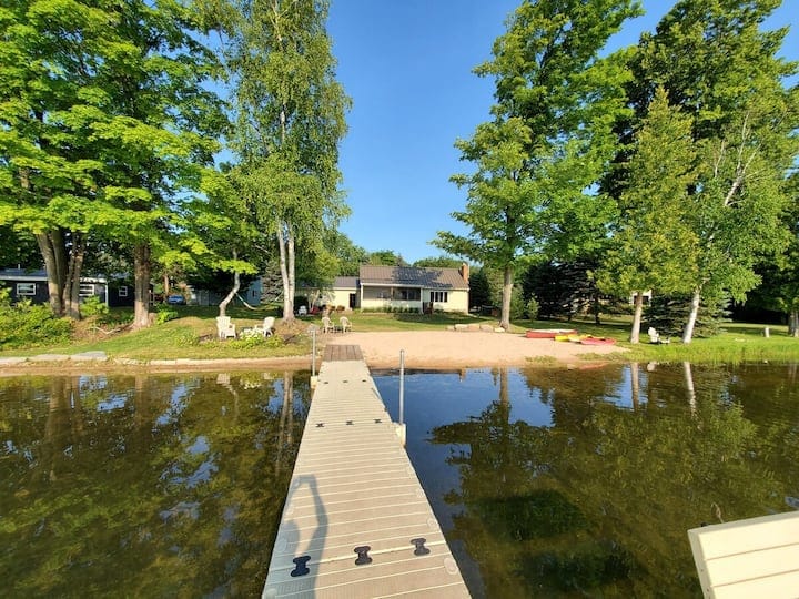 A spacious cottage is visible from the dock, set against a backdrop of lush green trees and clear blue skies. The sandy beach area is directly in front of the cottage, with calm lake waters reflecting its surroundings. Kayaks and yard games can be seen nearby.