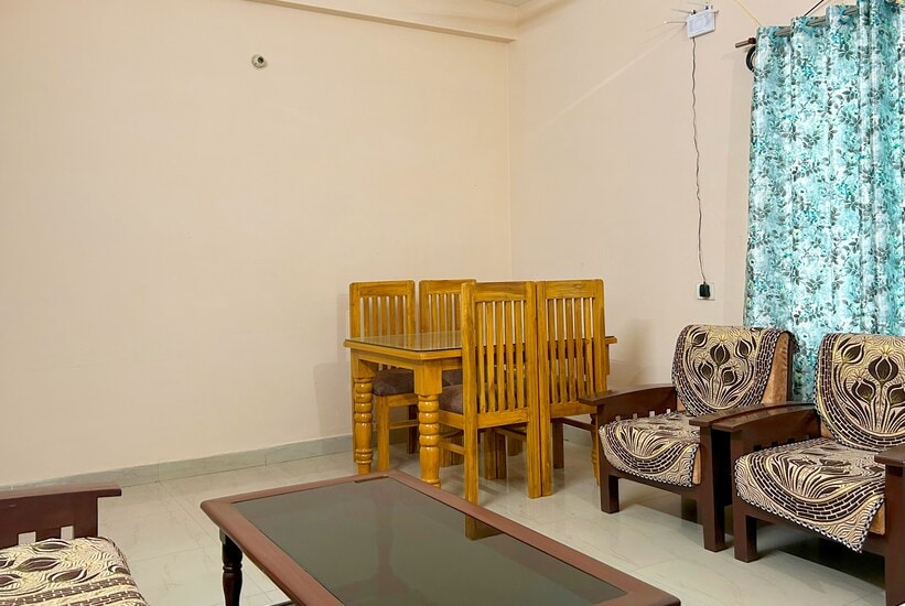 A living area features a wooden dining table accompanied by four matching chairs. Two upholstered armchairs with an intricate pattern are arranged around a dark wooden coffee table. Natural light enters through a window dressed with a floral curtain.