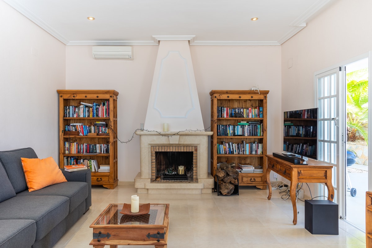 A spacious living area features comfortable seating on a gray sofa, complemented by orange accent pillows. Wooden bookshelves filled with books frame a decorative fireplace, while a television sits on a wooden stand. Natural light enters through glass doors, enhancing the room's inviting atmosphere.