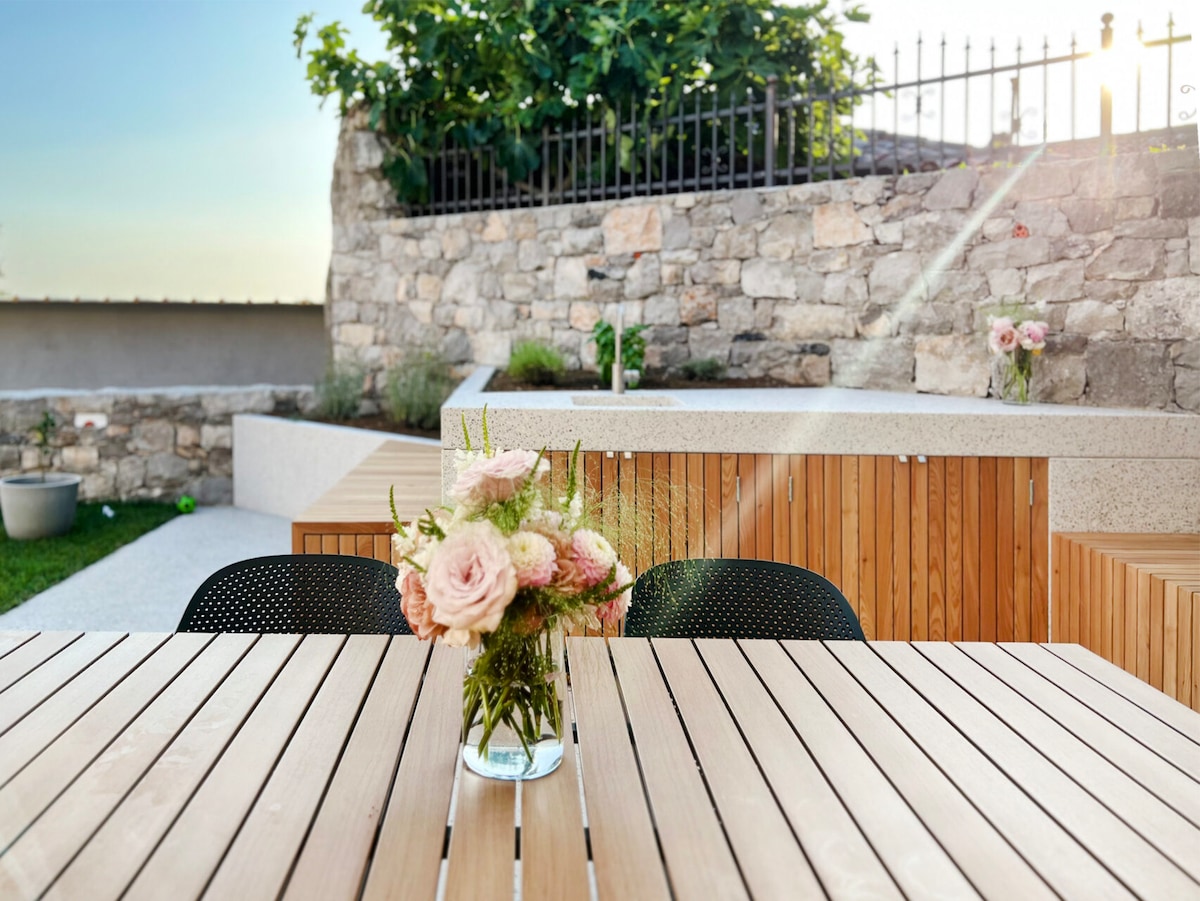 A wooden dining table is set outdoors, adorned with a vase of soft pink flowers. Surrounding benches are visible, with a stone wall in the background and greenery lining the raised garden beds. The setting is illuminated by warm sunlight.